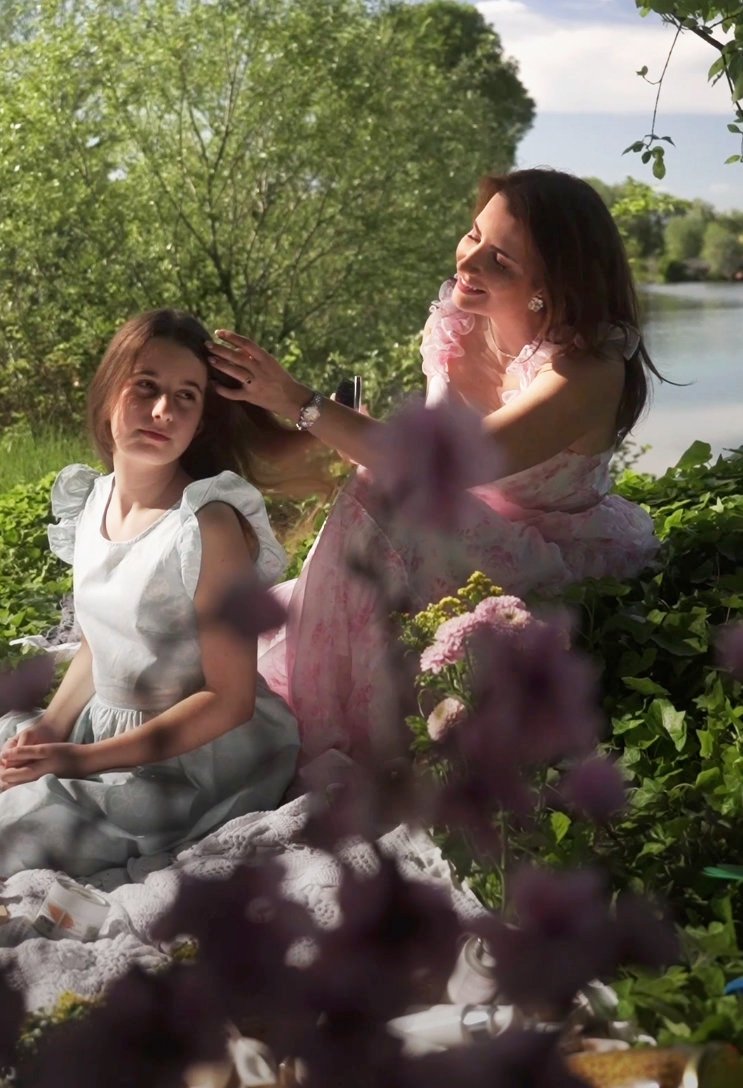 Vrouw en meisje zittend tussen bomen en bloemen aan het water.