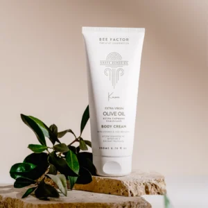 Witte tube Bee Factor-bodycrème met olijfolie naast een olijftak op lichte stenen.
