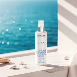 Een witte sprayflacon Essence of Mykonos van Bee Factor bij een raam met uitzicht op zee en kiezels.