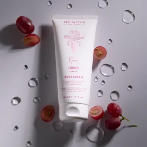 Witte tube Bee Factor-bodycrème met druiven tussen rode druiven en heldere druppels op een grijze achtergrond.