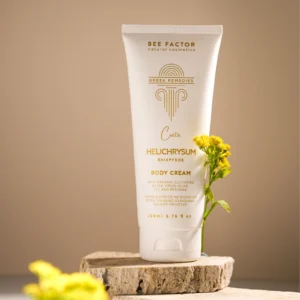 Witte tube Bee Factor-bodycrème met helichrysum naast gele bloemen op een houten schijf.